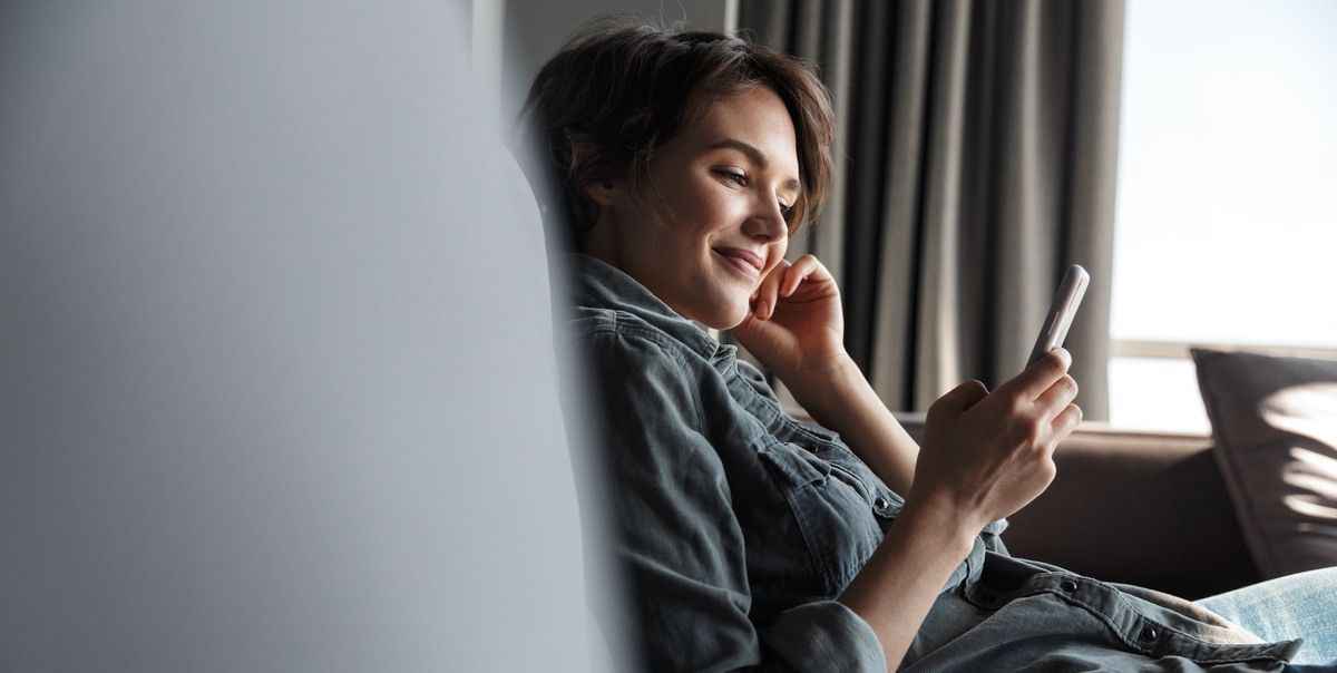 Image of nice young happy woman using mobile phone and smiling while sitting on sofa at living room